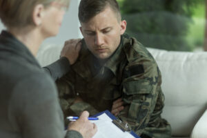 Concerned and upset young military man in uniform fatigues in female doctor's office. They're both seated, facing each other, and her hand is on his shoulder.