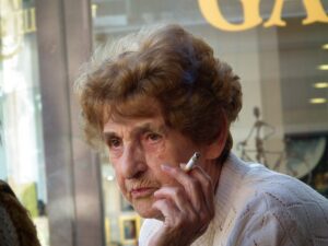 Elderly wrinkled woman with pursed lips and short attractive hairstyle, wearing a white sweater, holds an almost completely smoked cigarette.